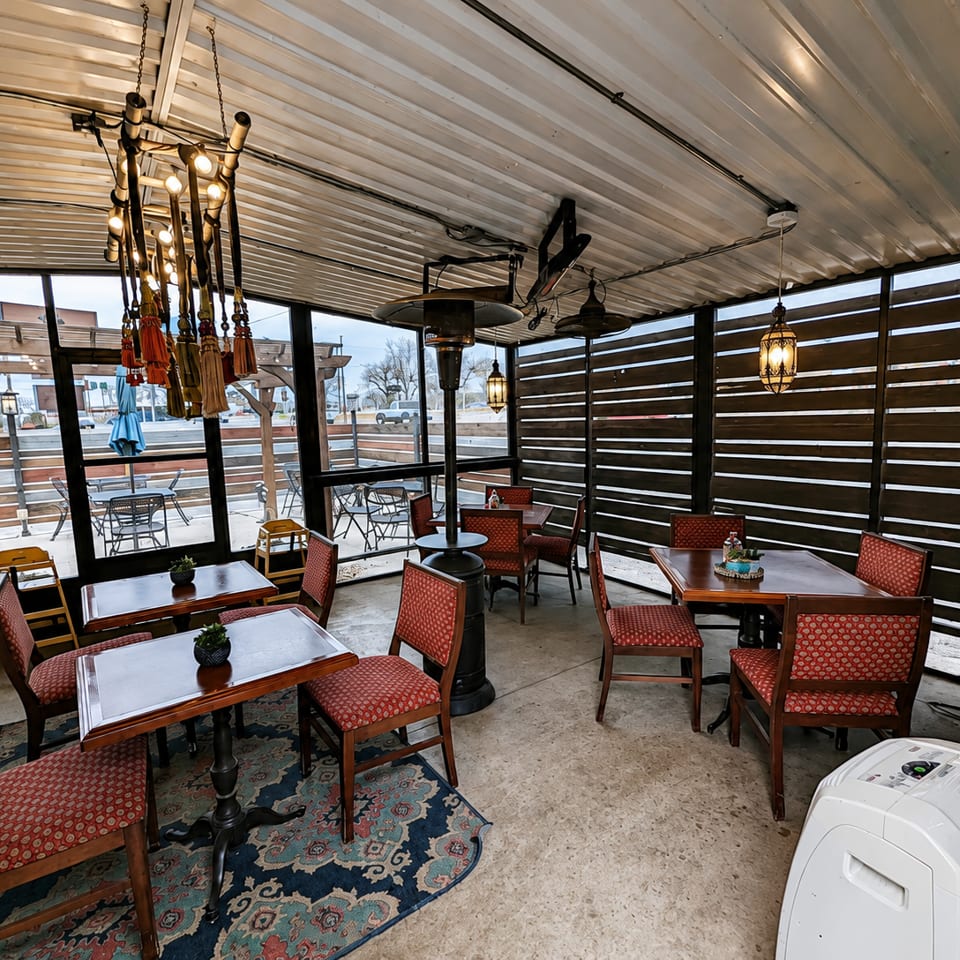 Enclosed restaurant patio with patterned red chairs, wooden tables, hanging lanterns, heaters, and outdoor seating visible through windows.