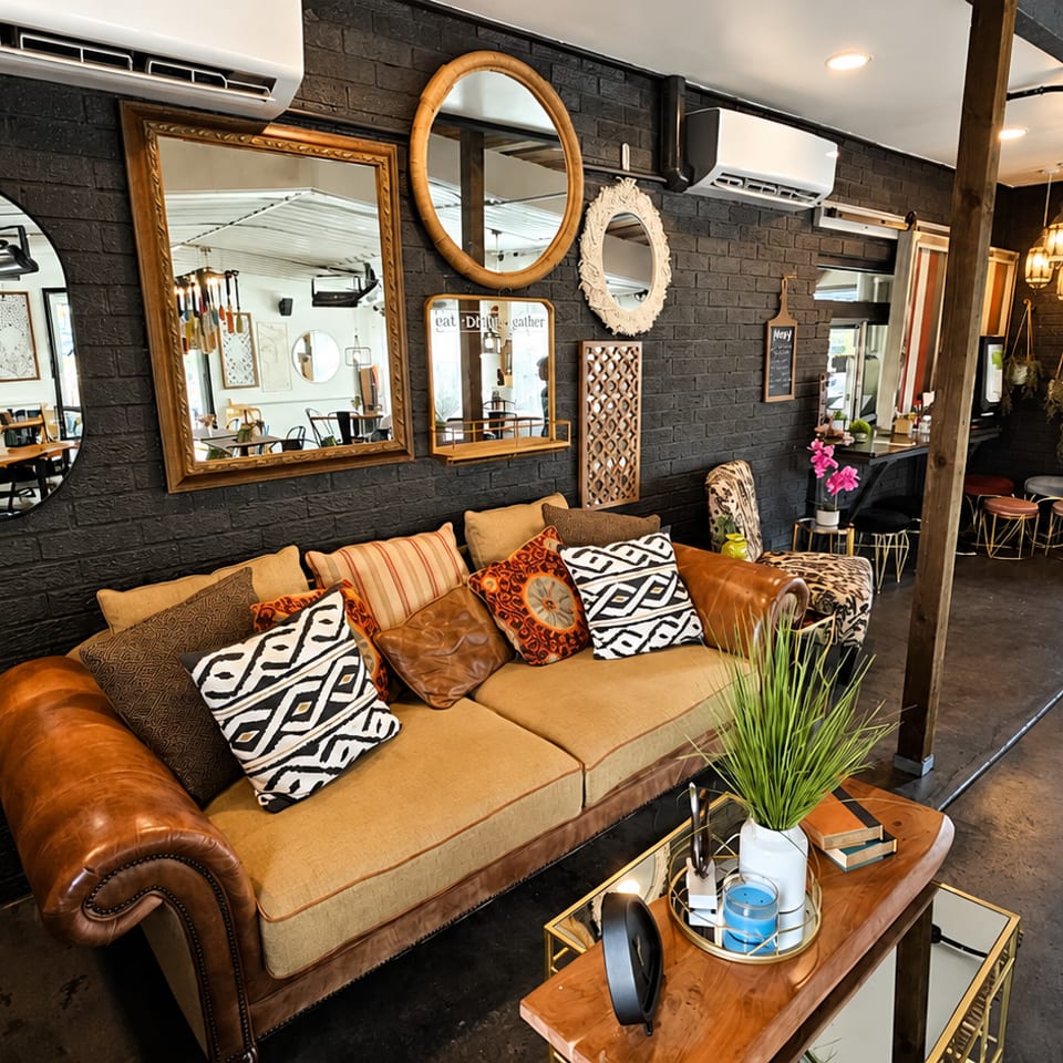 Cozy restaurant lounge with a tan leather sofa, patterned pillows, black brick walls, mirrors, plants, and pendant lighting.
