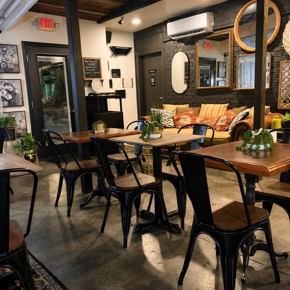 Cozy restaurant dining area with wooden tables, black metal chairs, plants, patterned pillows, and vintage mirrors.