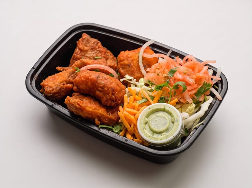 Fried chicken pieces with pickled onions, shredded carrots, cabbage, and green sauce in a black container.